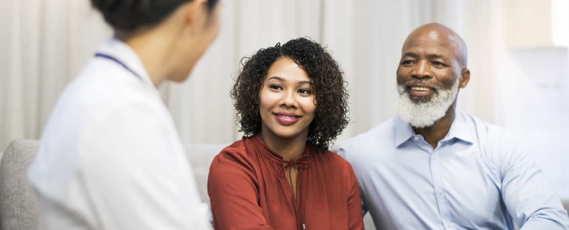 An African-American woman living with primary immunodeficiency and a bearded African-American man sit on a couch, smiling as they speak to a female doctor