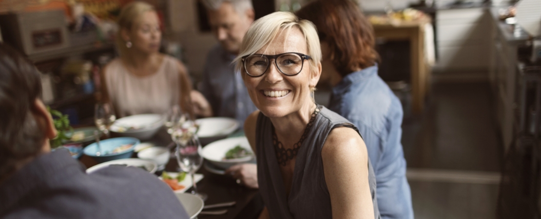 Smiling woman with short blond hair and glasses living with primary immunodeficiency sits at restaurant table with a group of friends.
