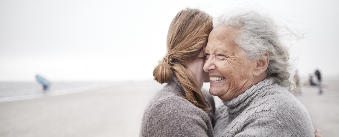 Senior woman smiles as she hugs younger woman on beach living with primary immunodeficiency