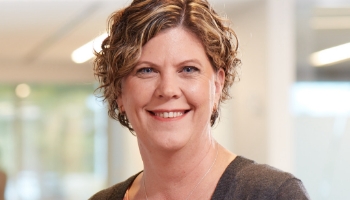 Patient Advocate Dayna, a smiling woman with brown hair standing in office, four children with PI