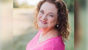 Patient Advocate Laura, a smiling woman with curly red hair and bright pink sweater standing in yard, both she and her son are diagnosed with PI